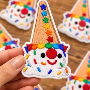 May include: An embroidered patch shaped like a clown face with a tan cone hat. The hat features a vertical row of colorful stars. The clown has a white face, a large red nose, black eyes, a red smile, and is decorated with colorful sprinkles.