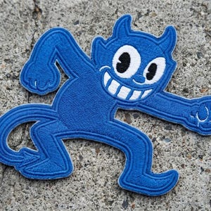 May include: A blue embroidered patch featuring a cartoon devil character with a wide, toothy grin, large white eyes with black pupils, two small horns, and a pointed tail, depicted in a dynamic, running pose with arms and legs extended.
