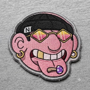 May include: An embroidered patch featuring a pink cartoon face wearing a black beanie with the letter N, gold hoop earrings, and yellow-tinted glasses. The character has a gold tooth and a purple mushroom-shaped object resting on its tongue.