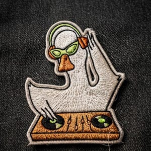 May include: An embroidered patch featuring a white goose wearing lime green headphones and matching sunglasses. The goose is positioned behind a brown DJ mixing console with two black vinyl records and vertical sliders, creating a music scene.