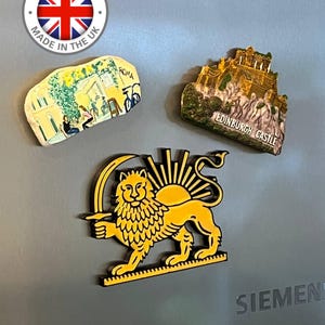 Könnte beinhalten: Drei Souvenir-Magnete auf einer grauen Oberfläche. Ein Magnet zeigt eine Szene aus Rom, Italien. Ein anderer zeigt Edinburgh Castle. Der dritte zeigt einen gelben Löwen mit Sonnenstrahlen. Ein "Made in the UK"-Aufkleber befindet sich oben links.