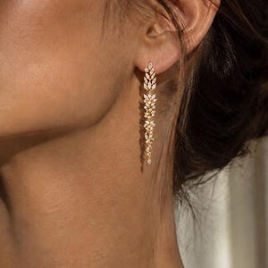 Könnte beinhalten: Ein langer, goldfarbener Ohrhänger mit einer vertikalen Anordnung von klaren, marquise-geschliffenen Steinen. Das Design verjüngt sich nach unten und erzeugt eine zarte, schlanke Silhouette, die mit einer polierten Metalloberfläche am Ohrläppchen hängt.