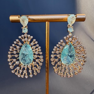 May include: A pair of gold-toned earrings with a light blue teardrop-shaped gemstone at the center. The earrings feature a sunburst design with small, clear gemstones and a smaller light blue gemstone at the top.