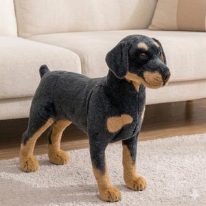 Op de afbeelding: Een zwart en bruin pluche hondenspeelgoed. De hond heeft bruine poten, borst en gezicht. Het speelgoed staat op een wit tapijt voor een beige bank. De hond heeft hangende oren en een korte staart.