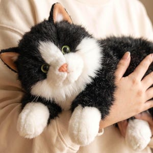 Può includere: Un peluche a forma di gatto con pelliccia bianca e nera, occhi verdi, naso rosa e zampe bianche. Il giocattolo ha orecchie a punta con interno color marrone chiaro e una consistenza soffice, che riproduce un gatto tuxedo in posizione seduta.