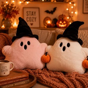 May include: Two plush ghost-shaped cushions, one light pink and one white, each wearing a black witch hat. The white ghost holds two small orange plush pumpkins. Both ghosts feature embroidered black oval eyes and an open mouth expression.