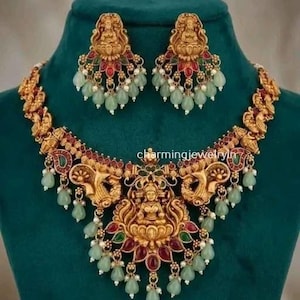 Op de afbeelding: Een goudkleurige ketting en bijpassende oorbellen met een centrale godheidsfiguur op een lotus, geaccentueerd met rode en groene stenen, kleine witte parels en bungelende lichtgroene druppelvormige kralen. De tekst charmingjewelryin is zichtbaar.