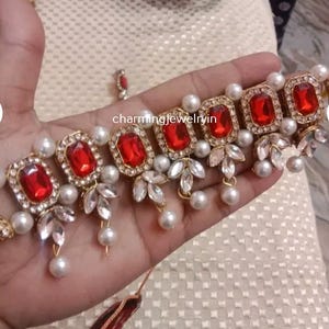 Könnte beinhalten: Ein goldfarbenes Choker-Halsband mit roten rechteckigen Edelsteinen, umgeben von klaren Kristallen und weißen Perlen. Die Halskette hat ein Blumendesign mit hängenden Perlen. Der Text "charmingjewelryin" ist sichtbar.