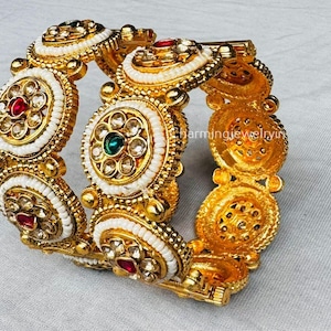 Peut inclure: Un bracelet jonc doré avec des détails complexes. Le bracelet présente des accents de perles blanches et des motifs circulaires avec des pierres précieuses rouges, vertes et transparentes. Le fermoir est visible sur le côté droit.