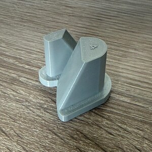 Può includere: Un componente in plastica grigia stampato in 3D caratterizzato da due torri verticali distinte con cime inclinate, ciascuna montata su una base piatta. La torre più grande presenta un piccolo numero 4 in rilievo sulla superficie superiore e una trama.