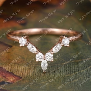 May include: A rose gold ring with a curved design, set with multiple clear gemstones. The ring features a series of round and marquise-cut stones, creating a delicate and elegant appearance. The ring is designed to be worn as a wedding band or fashion ring.