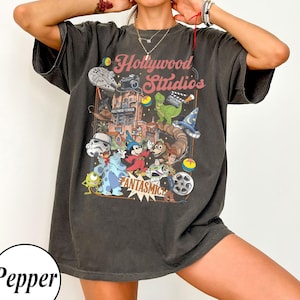 Peut inclure: Un t-shirt gris anthracite à manches courtes avec une impression graphique portant le texte Hollywood Studios et Fantasmic. Le motif comprend divers personnages animés, une bobine de film, un appareil photo et un vaisseau spatial en couleur.