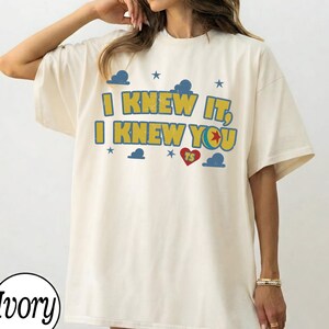 May include: An ivory short-sleeve t-shirt featuring the yellow text I KNEW IT, I KNEW YOU with a red heart containing the letters TS, surrounded by blue cloud and star graphics. The design has a distressed, weathered appearance on the fabric.