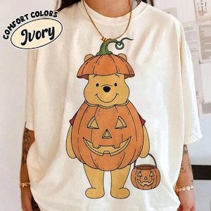 May include: An ivory t-shirt featuring a graphic of a yellow bear character wearing a pumpkin costume and holding a small pumpkin-shaped bucket. The text COMFORT COLORS IVORY is printed in a black oval in the upper left corner of the shirt.