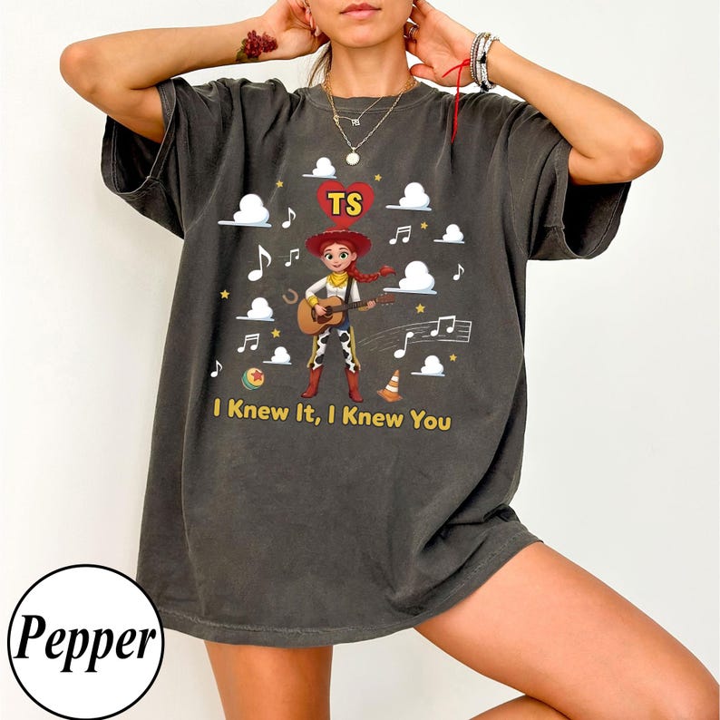 May include: A charcoal grey short-sleeve t-shirt featuring a graphic of the character Jessie from Toy Story playing a guitar. The design includes a red heart with the letters TS, white clouds, musical notes, and the text I Knew It, I Knew You.
