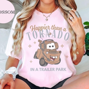 May include: Light pink t-shirt with the text "Happier than a Tornado in a Trailer Park" and a cartoon image of a brown tow truck. The word "Blossom" is in a pink circle in the upper left.