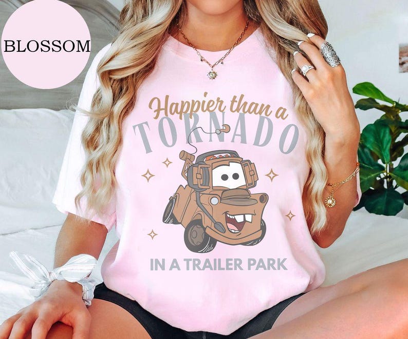May include: Light pink t-shirt with the text "Happier than a Tornado in a Trailer Park" and a cartoon image of a brown tow truck. The word "Blossom" is in a pink circle in the upper left.