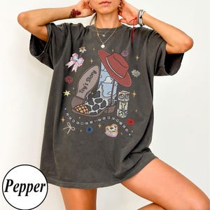 Op de afbeelding: Een antracietgrijs t-shirt met korte mouwen en een grafische print van een cowboylaars met een koeienprint neus, een rode hoed en blokken met de tekst Toy's Story. Het ontwerp bevat kleine sterretjes, bloemen en een strik.