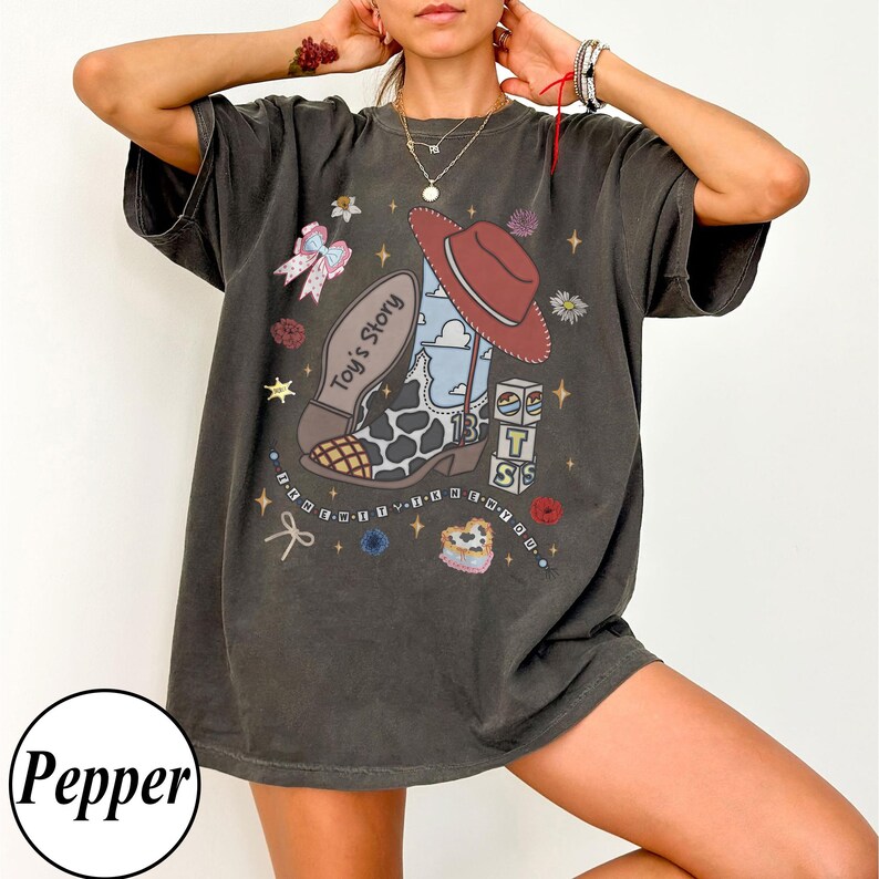Op de afbeelding: Een antracietgrijs t-shirt met korte mouwen en een grafische print van een cowboylaars met een koeienprint neus, een rode hoed en blokken met de tekst Toy's Story. Het ontwerp bevat kleine sterretjes, bloemen en een strik.
