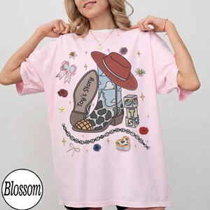 Op de afbeelding: Een lichtroze t-shirt met een grafische print van een cowboylaars met koeienvlekkenpatroon, een rode hoed en de tekst Toy's Story. Het ontwerp bevat kleine illustraties van een strik, alfabetblokken, bloemen en een kralensnoer met tekst.