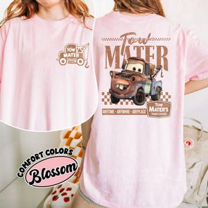 May include: Pink Comfort Colors t-shirt featuring a brown graphic of Tow Mater from the movie Cars. The front has a small graphic of Mater's tow truck, and the back displays a larger image with the text "Tow Mater".