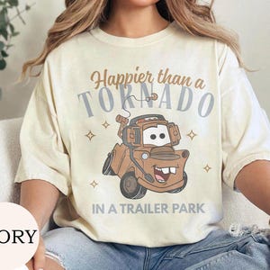 May include: Ivory-colored t-shirt with the text "Happier than a Tornado in a Trailer Park" and a cartoon image of a brown tow truck. The design includes stars and is printed on a short-sleeved shirt.