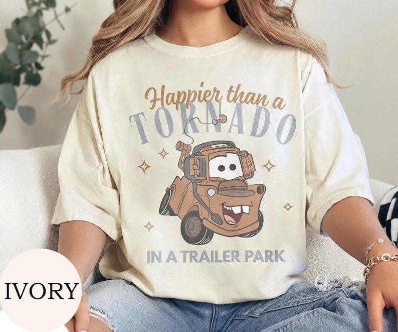 May include: Ivory-colored t-shirt with the text "Happier than a Tornado in a Trailer Park" and a cartoon image of a brown tow truck. The design includes stars and is printed on a short-sleeved shirt.