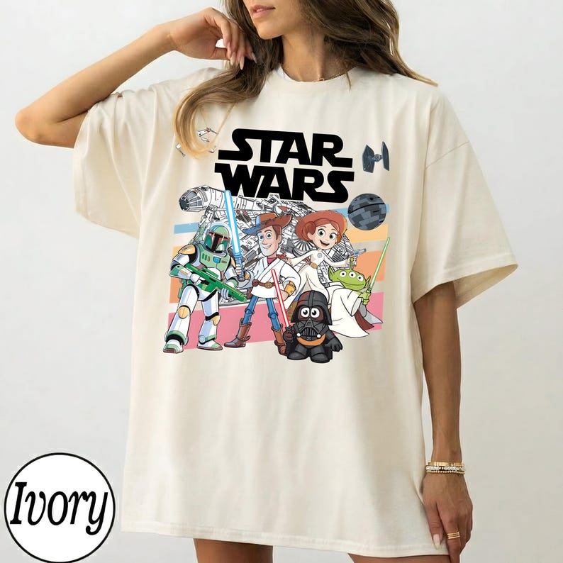 May include: Ivory-coloured t-shirt with a colourful graphic of Star Wars characters. The design includes the Star Wars logo and illustrations of characters like Darth Vader, Woody, and Buzz Lightyear. The shirt has short sleeves and a crew neck.