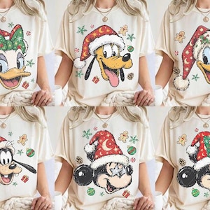 Könnte beinhalten: Eine Kollektion von sechs cremefarbenen T-Shirts, die jeweils eine andere Disney-Figur mit einer festlichen Weihnachtsmütze zeigen. Die Designs umfassen Daisy Duck, Pluto, Donald Duck, Goofy und zwei Varianten von Mickey Mouse, umgeben von kleinen Lebkuchenmännern, Tannenzapfen und Weihnachtskugeln.