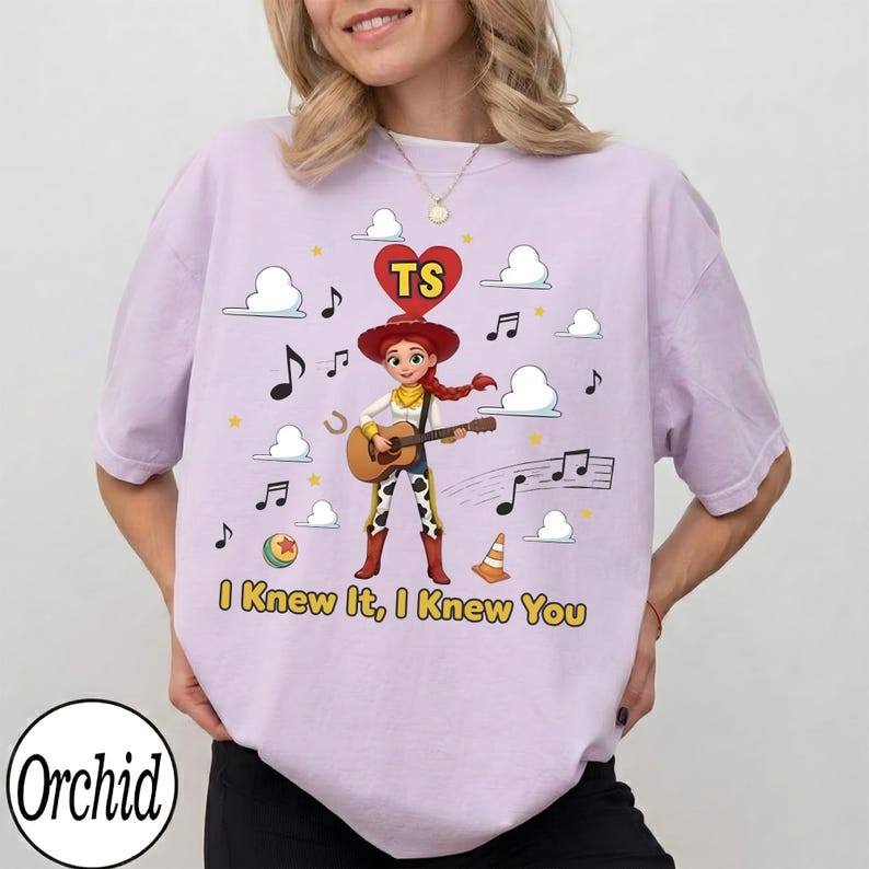 May include: A light purple t-shirt featuring a graphic of a cartoon cowgirl playing an acoustic guitar. Above her is a red heart with the letters TS. The design includes musical notes, clouds, a ball, and a traffic cone, with the text I Knew It, I Knew You.