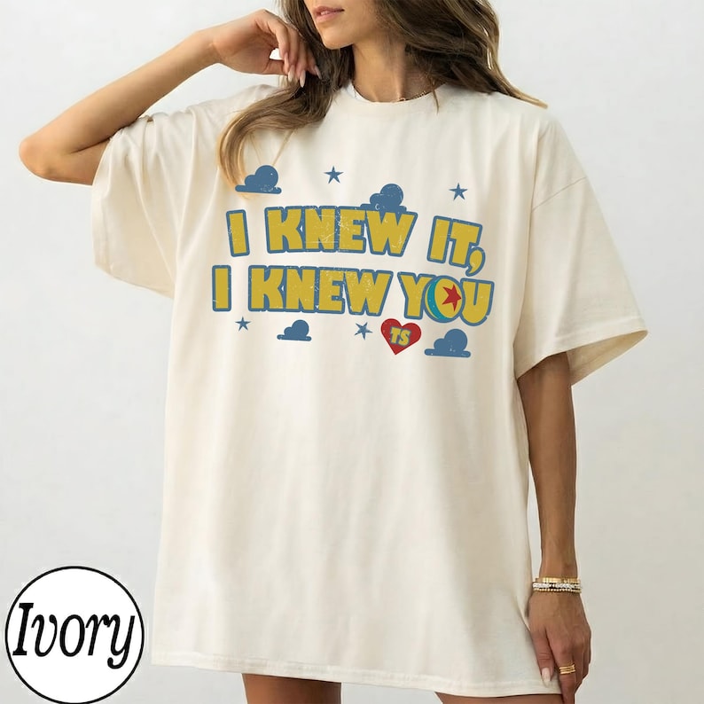 May include: An ivory short-sleeve t-shirt featuring the yellow text I KNEW IT, I KNEW YOU with a red heart containing the letters TS, surrounded by blue cloud and star graphics. The design has a distressed, weathered appearance on the fabric.