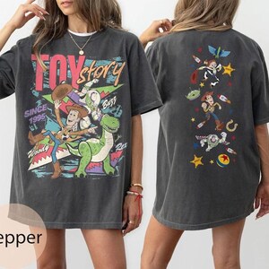 Puede incluir: Una camiseta gris carb&oacute;n con gr&aacute;ficos de Toy Story. El frente muestra a Woody, Buzz Lightyear y Rex con el texto "TOY story SINCE 1995". La parte posterior muestra una disposici&oacute;n vertical de personajes que incluye a Woody, Buzz y un alien con estrellas.