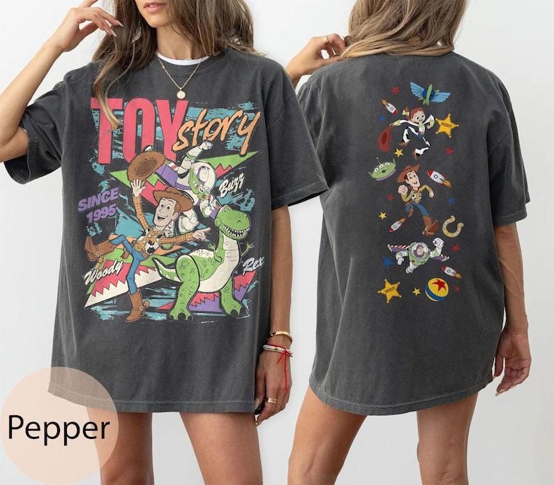 Puede incluir: Una camiseta gris carb&oacute;n con gr&aacute;ficos de Toy Story. El frente muestra a Woody, Buzz Lightyear y Rex con el texto "TOY story SINCE 1995". La parte posterior muestra una disposici&oacute;n vertical de personajes que incluye a Woody, Buzz y un alien con estrellas.