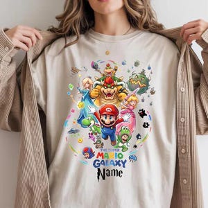 May include: A beige t-shirt with a vibrant graphic of Super Mario Galaxy characters. The design showcases Mario, Princess Peach, Bowser, and others, with the text "THE SUPER MARIO GALAXY Name". The shirt is a neutral colour.