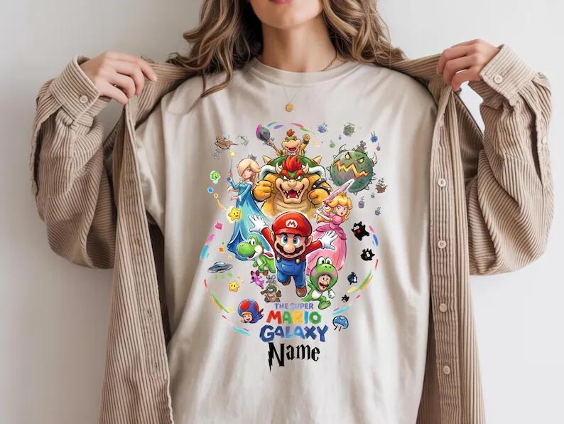May include: A beige t-shirt with a vibrant graphic of Super Mario Galaxy characters. The design showcases Mario, Princess Peach, Bowser, and others, with the text "THE SUPER MARIO GALAXY Name". The shirt is a neutral colour.