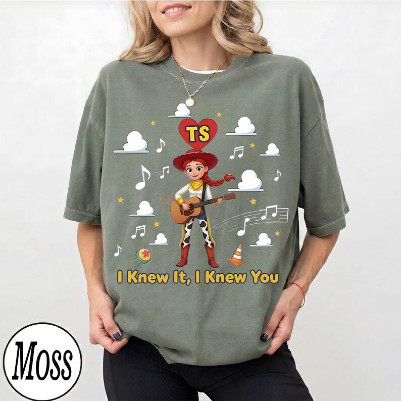 May include: An olive green crewneck t-shirt featuring a graphic of a cowgirl character playing an acoustic guitar. Above the character is a red heart with the letters TS. The text I Knew It, I Knew You is printed below, surrounded by clouds, stars, and musical notes.