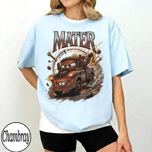Puede incluir: Camiseta azul claro con una ilustraci&oacute;n de Mater de la pel&iacute;cula Cars. El dise&ntilde;o presenta la palabra "MATER" en marr&oacute;n sobre un personaje de cami&oacute;n de remolque marr&oacute;n. El gr&aacute;fico incluye herramientas y humo alrededor del cami&oacute;n.