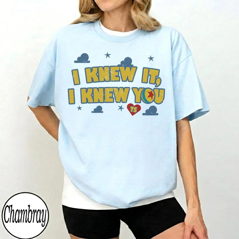 May include: A light blue t-shirt featuring the yellow text I KNEW IT, I KNEW YOU with a red heart containing the letters TS, surrounded by small blue cloud and star graphics. The design has a distressed, vintage-inspired texture.
