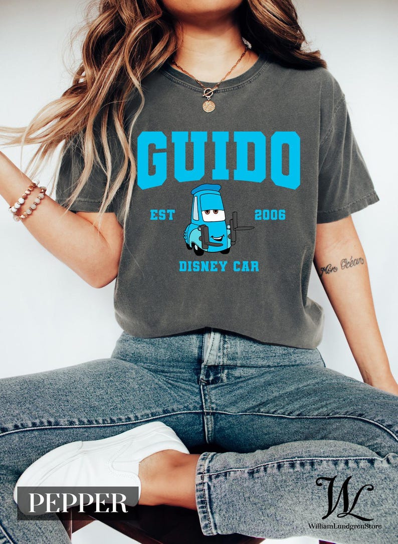 Puede incluir: Camiseta gris oscuro con el nombre "Guido" en letras grandes de color azul y una imagen de dibujos animados de un coche Disney azul. El texto "EST 2006 DISNEY CAR" tambi&eacute;n est&aacute; impreso en la camiseta. La camiseta es de estilo casual.