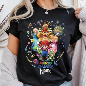May include: Black t-shirt with a vibrant graphic of Super Mario Galaxy characters, including Mario, Princess Peach, and Bowser. The design is surrounded by stars and planets. The text on the shirt reads "The Super Mario Galaxy Name".