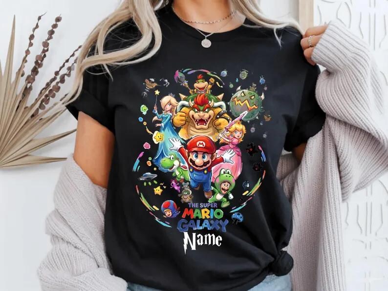 May include: Black t-shirt with a vibrant graphic of Super Mario Galaxy characters, including Mario, Princess Peach, and Bowser. The design is surrounded by stars and planets. The text on the shirt reads "The Super Mario Galaxy Name".