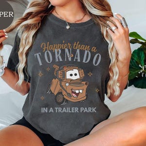 May include: Dark gray t-shirt featuring a cartoon image of Mater from the movie Cars. The shirt has the text "Happier than a Tornado in a Trailer Park" in brown and gray lettering. The design includes small star accents.