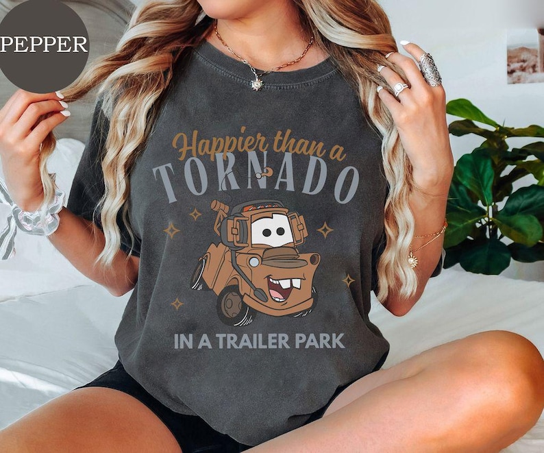 May include: Dark gray t-shirt featuring a cartoon image of Mater from the movie Cars. The shirt has the text "Happier than a Tornado in a Trailer Park" in brown and gray lettering. The design includes small star accents.