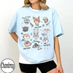 Może przedstawiać: Jasnoniebieska koszulka z kresk&oacute;wkowymi ilustracjami postaci z filmu Zwierzogr&oacute;d, w tym Nicka Wilde'a, Judy Hopps i Flasha. Koszulka ma imiona postaci i gwiazdki. Nazwa marki "Chambray" znajduje się w lewym dolnym rogu.