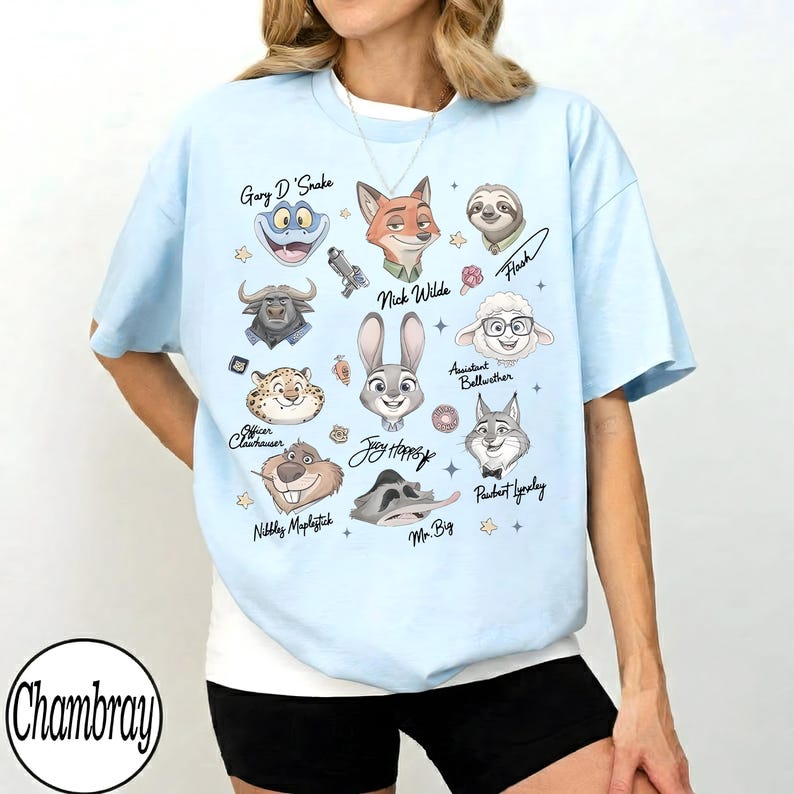 Może przedstawiać: Jasnoniebieska koszulka z kresk&oacute;wkowymi ilustracjami postaci z filmu Zwierzogr&oacute;d, w tym Nicka Wilde'a, Judy Hopps i Flasha. Koszulka ma imiona postaci i gwiazdki. Nazwa marki "Chambray" znajduje się w lewym dolnym rogu.