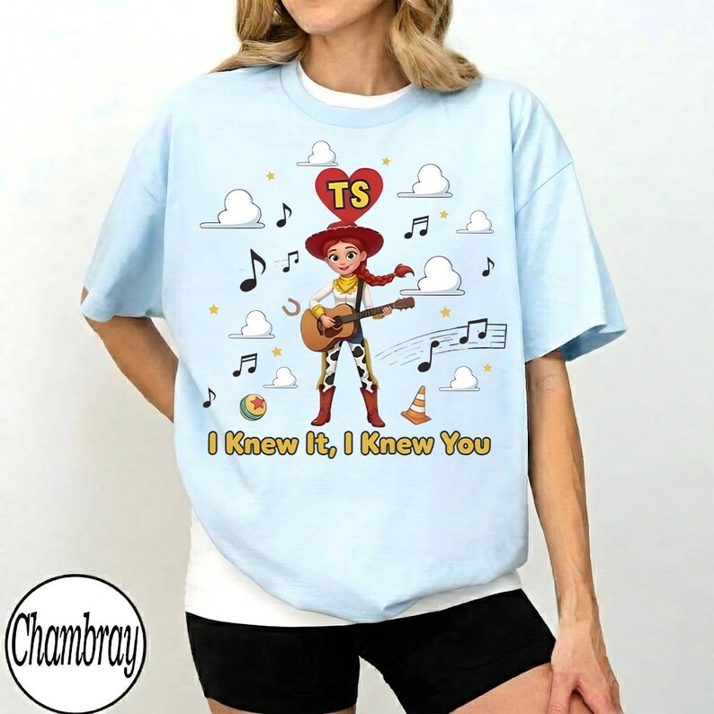 May include: A light blue t-shirt featuring a graphic of a cowgirl character playing an acoustic guitar. Above the character is a red heart with the letters TS. The design includes white clouds, musical notes, a ball, and a traffic cone. The text "I Knew It, I Knew You" is printed in yellow at the bottom.