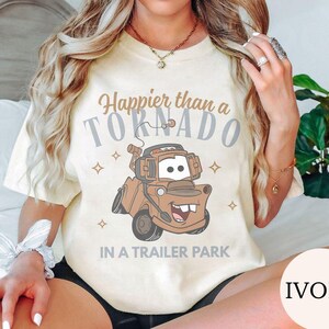 May include: Ivory-colored t-shirt featuring a cartoon image of Tow Mater from the movie Cars. The shirt has the text "Happier than a Tornado in a Trailer Park" in brown and gray lettering. The design includes stars.