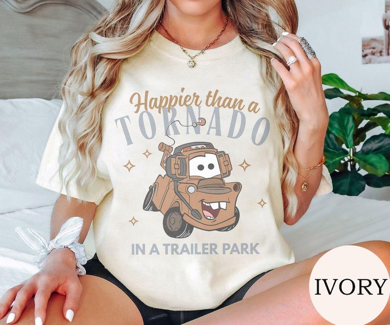 May include: Ivory-colored t-shirt featuring a cartoon image of Tow Mater from the movie Cars. The shirt has the text "Happier than a Tornado in a Trailer Park" in brown and gray lettering. The design includes stars.