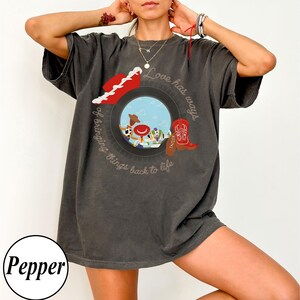 Op de afbeelding: Een antracietgrijs t-shirt met korte mouwen en een cirkelvormige afbeelding met een blauwe lucht, stripfiguren, een rode cowboyhoed en een rode cowboylaars. De tekst luidt Love has ways of bringing things back to life.