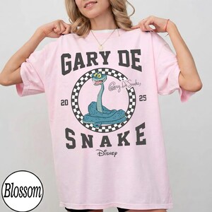 May include: Light pink t-shirt with a graphic of a blue snake inside a black and white checkered circle. The text "GARY DE SNAKE" is above and below the snake graphic, with "Disney" below. The numbers "20" and "25" flank the circle.